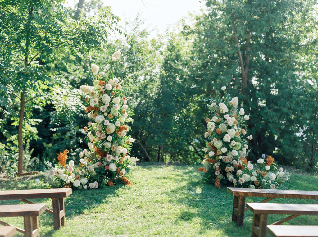 A ceremony set up with an asymmetrical arch and wooden benches lining the aisle in a grassy field with trees